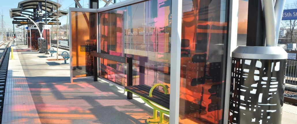 Sharon Dowell's glass windscreen with laminated black, pink, & orange patterns & human silhouettes as through a passing train window, cast colorful shadows on the 25th St Station light rail platform. Canopy columns with b&w metal abstract designs complement the windscreen.