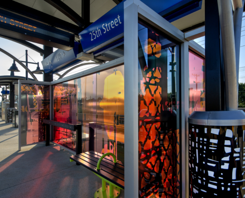 Glass windscreen with laminated black, pink, & orange patterns & human silhouettes as through a passing train window cast colorful shadows on the 25th St Station light rail platform. Canopy columns with b&w metal abstract designs complement the windscreen.