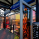 Glass windscreen with laminated black, pink, & orange patterns & human silhouettes as through a passing train window cast colorful shadows on the 25th St Station light rail platform. Canopy columns with b&w metal abstract designs complement the windscreen.