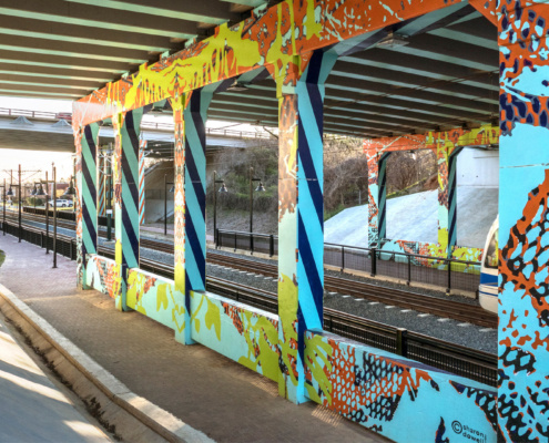 I-277 & 11th St underpass over light rail track & walkway in Charlotte. Painted in sky blue with cobalt blue & orange diagonal, repeating, lined patterns down sides of concrete support ballasts. Mesh-like & organic patterns in various bright colors throughout.