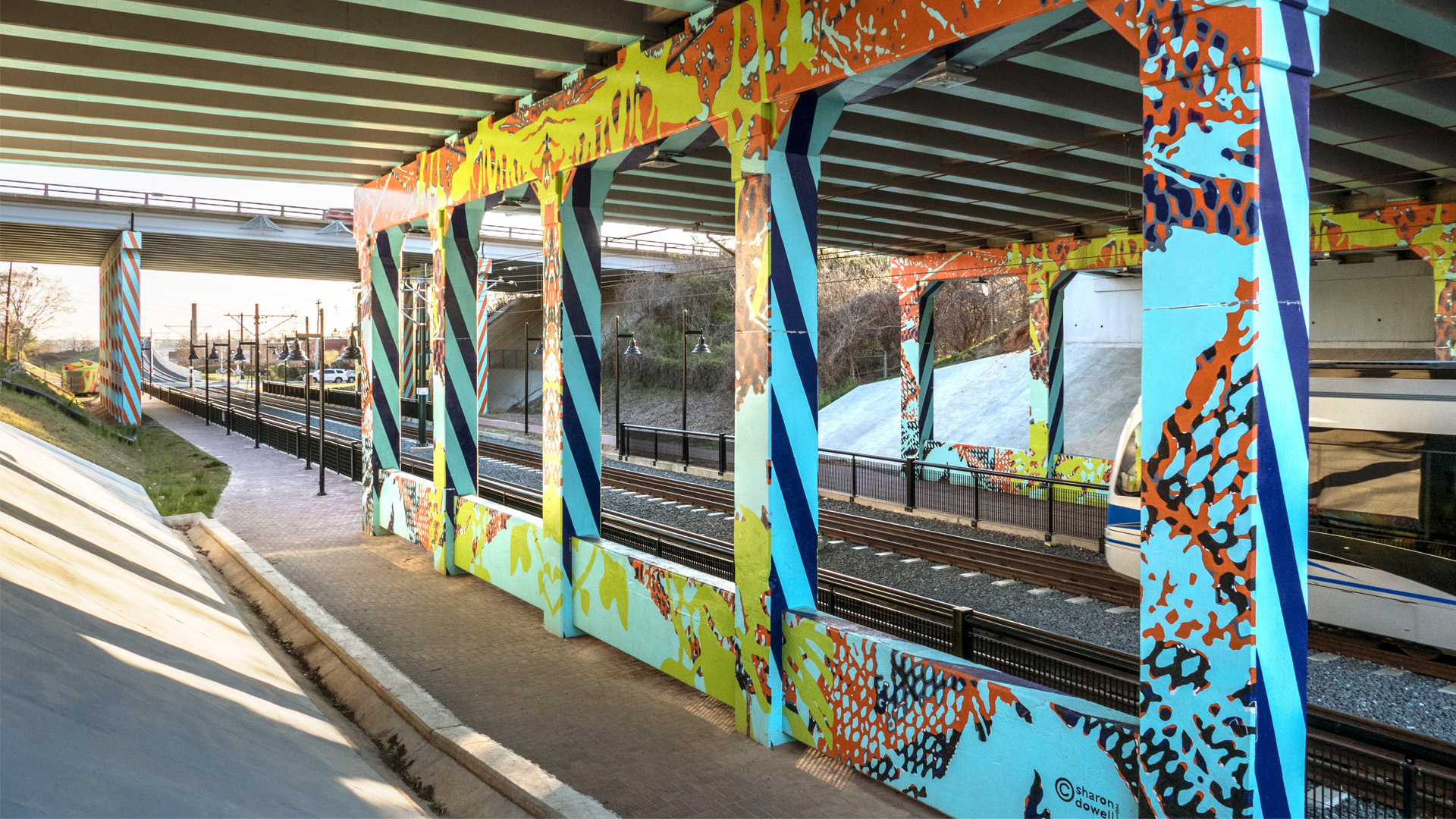 I-277 & 11th St underpass over light rail track & walkway in Charlotte. Painted in sky blue with cobalt blue & orange diagonal, repeating, lined patterns down sides of concrete support ballasts. Mesh-like & organic patterns in various bright colors throughout.