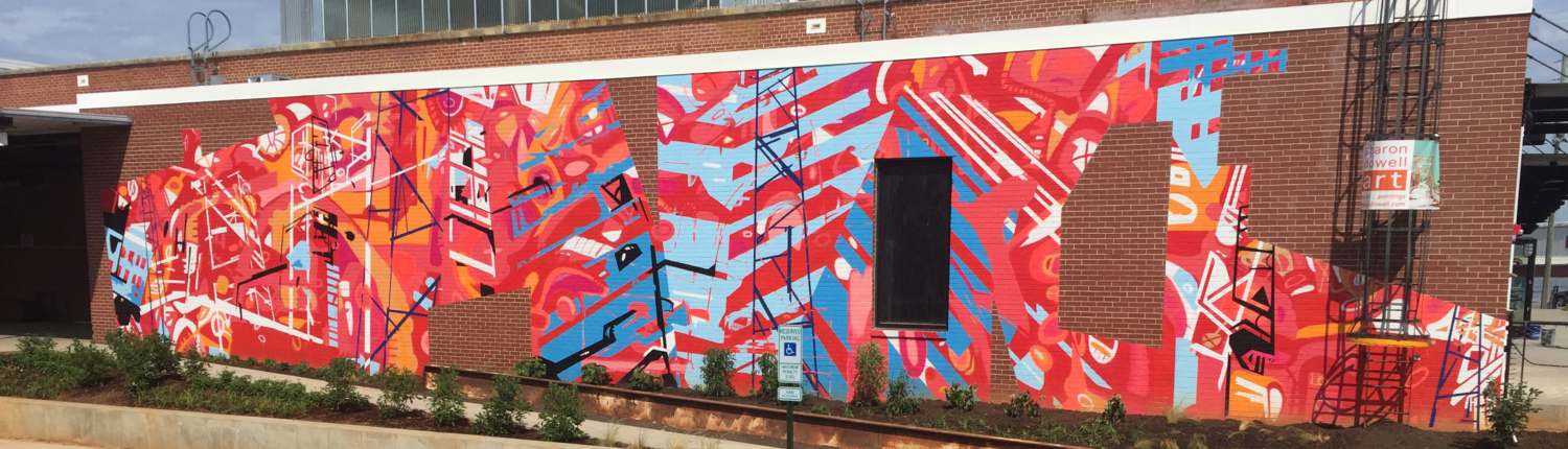 20’ h x 80’ w hand & spray-painted energetic, abstract mural with overlapping planes of reds, blues & oranges. Architectural lines in blues, black & white echo industrial buildings and towers of surroundings. Precise geometric shapes of exposed, unpainted red brick. Located on exterior wall of Goodyear Arts building in Charlotte’s Camp North End.