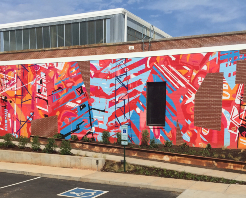 20’ h x 80’ w hand & spray-painted energetic, abstract mural with overlapping planes of reds, blues & oranges. Architectural lines in blues, black & white echo industrial buildings and towers of surroundings. Precise geometric shapes of exposed, unpainted red brick. Located on exterior wall of Goodyear Arts building in Charlotte’s Camp North End.