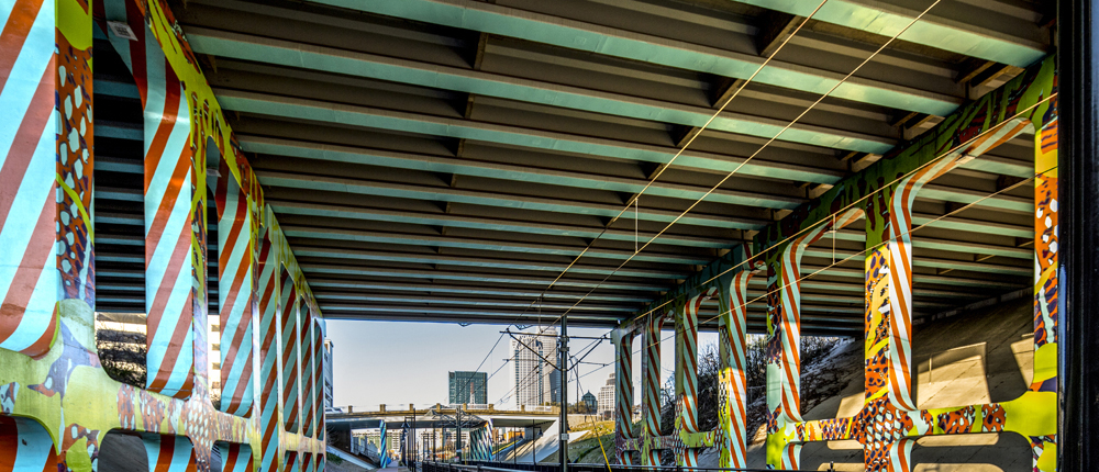 Detail of I-277 & 11th St underpass over light rail track & walkway in Charlotte. Sidewalk perspective looking down track towards city. Mural painted sky blue with cobalt blue & orange diagonal, repeating, lined patterns down sides of concrete support ballasts. Mesh-like & organic patterns in various bright colors throughout.