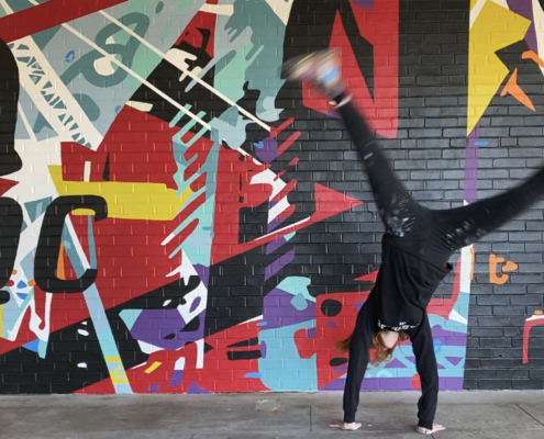 Figure in black clothing cartwheels in front of energetic mural of geometric shapes & abstracted trusses & beams in bold colors on brick wall. Charlotte NC’s crown logo and QC painted prominently in black on left side of mural. Located outside Bart’s Bottle Shop, Eastway Shopping Center.
