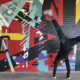 Figure in black clothing cartwheels in front of energetic mural of geometric shapes & abstracted trusses & beams in bold colors on brick wall. Charlotte NC’s crown logo and QC painted prominently in black on left side of mural. Located outside Bart’s Bottle Shop, Eastway Shopping Center.