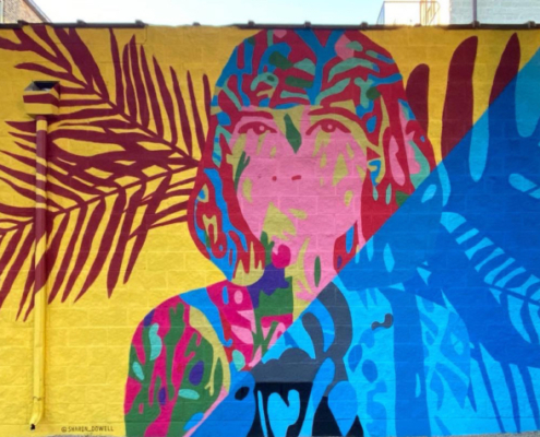 15” h x 24’ w mural on exterior concrete wall of woman painted in vibrant colors with shoulder-length hair facing forward, looking outward. Mural bisected diagonally: yellow background with maroon palm fronds on left side, right side blocked in all blues. In shadows of Ashland Ave L Train bridge, Lakeview Chicago.