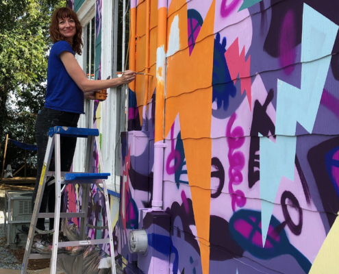Sharon Dowell painting a colorful, two-story mural of comic book-style lightning bolts & playful shapes & lines covering the entire exterior wall of Girls Rock CLT headquarters.