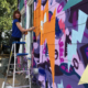 Sharon Dowell painting a colorful, two-story mural of comic book-style lightning bolts & playful shapes & lines covering the entire exterior wall of Girls Rock CLT headquarters.