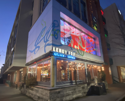 Corner view from street of 10’ h x 50’ w colorful, whimsical, abstract window film and building wrap over slow-changing, multi-colored LED lightbox. Above Leroy Fox in Charlotte’s Southend. Collaboration with Christopher Hoslton for I Heart Rail Trail 2021.